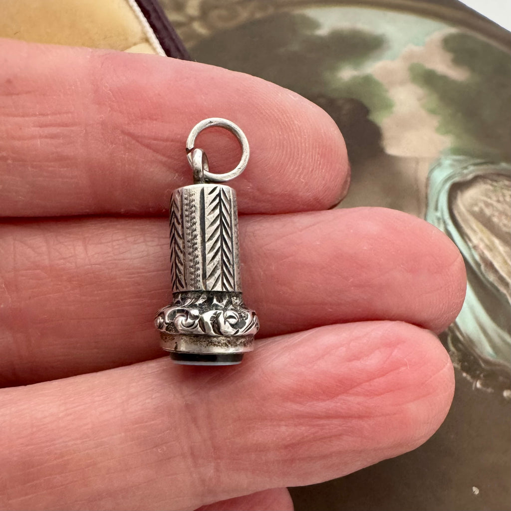 A small antique miniature silver wax seal fob charm or pendant with engraved geometric and floral details, held between fingers against a blurred background.