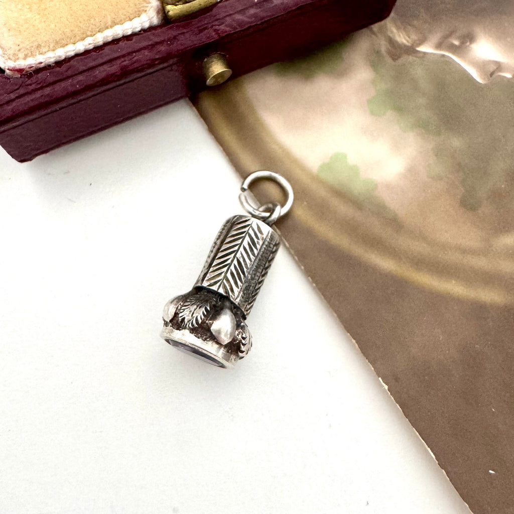 An antique miniature wax seal fob with engraved leaf-like detailing, shown on a light surface with a metallic gold-toned sheet and a dark wooden box in the background.
