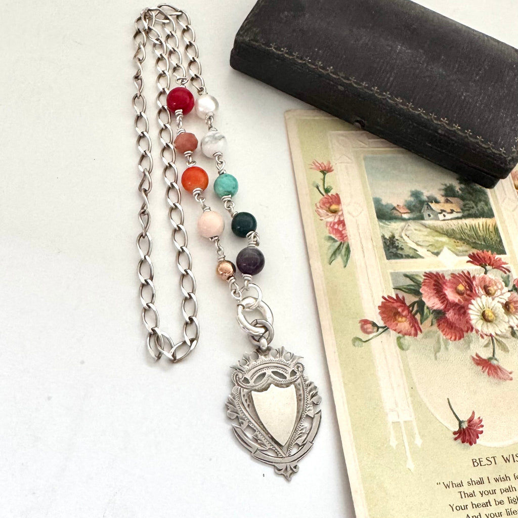 Necklace with multicolored beads and antique silver watch fob on a white surface with a jewelry box and floral card.