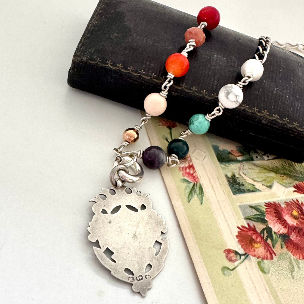 Necklace with multicolored beads and antique silver watch fob on a white surface with a jewelry box and floral card.
