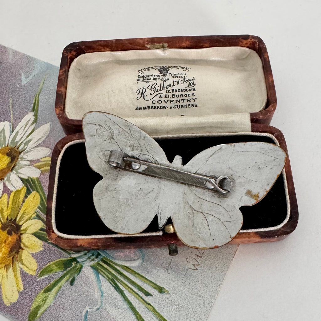 Back of Art Deco era butterfly-shaped brooch with a silver-toned metal pin back, displayed inside an open antique presentation box. The box has a brown wood-grain exterior and a cream-colored interior with printed branding for R.