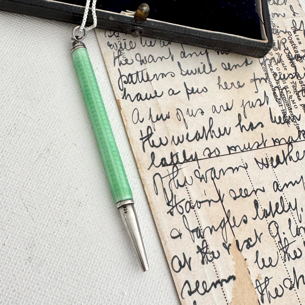 A close-up of an Art Deco silver and guilloche enamel propelling pencilresting on a textured white surface next to a vintage handwritten letter and a dark wooden box.