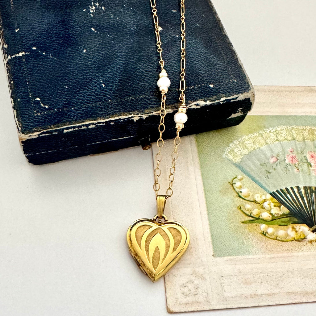 Gold heart-shaped necklace with pearl chain on a vintage box and fan background