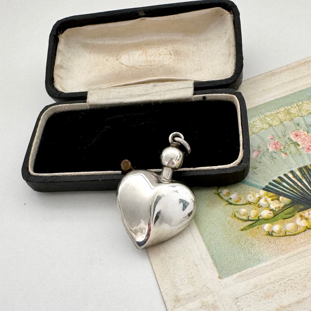 Silver heart-shaped perfume bottle pendant in an open black box with a vintage card in the background