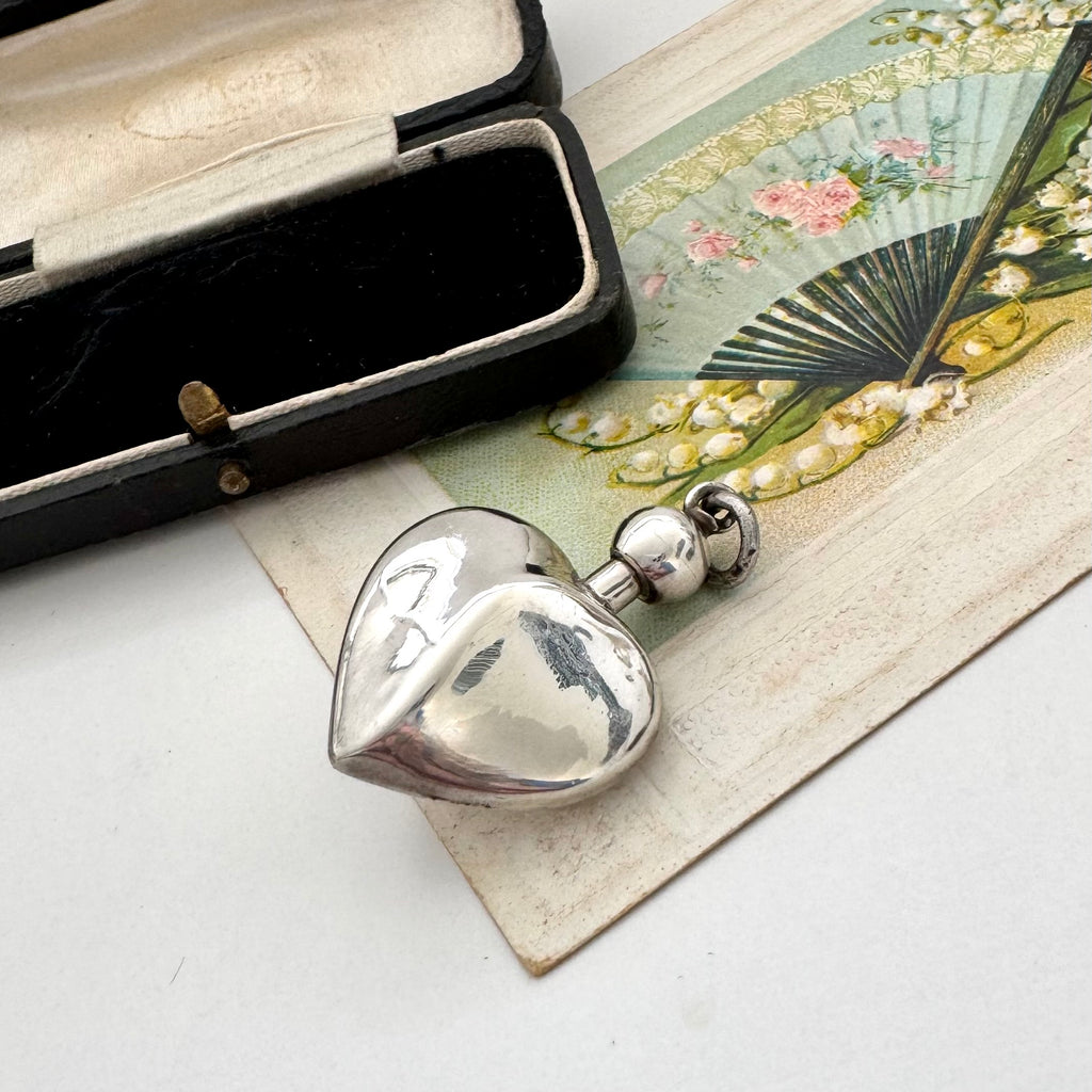 Silver heart-shaped perfume bottle pendant with an open black box with a vintage card in the background