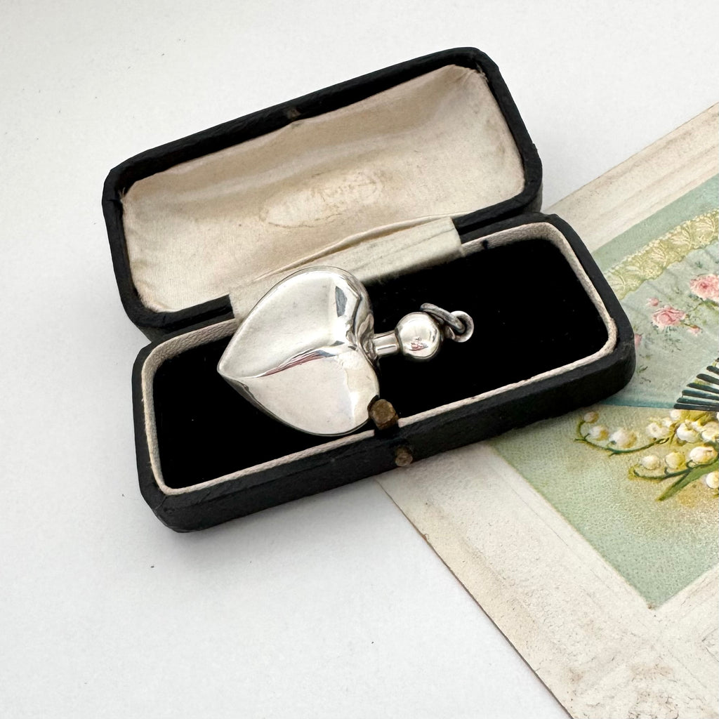 Silver heart-shaped perfume bottle pendant in an open black jewelry box on a white surface.