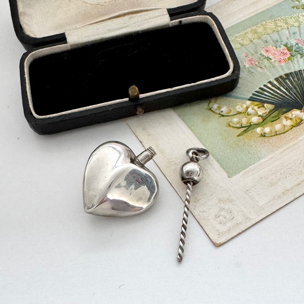 Silver heart-shaped perfume bottle pendant with an open black jewelry box on a white surface.