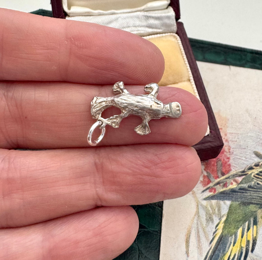 Silver platypus charm/pendant held between fingers with a decorative box in the background