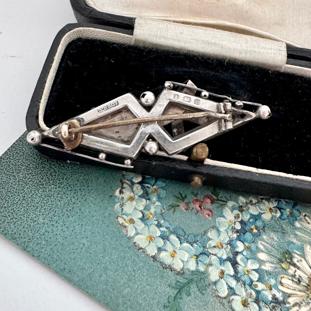 Back of Victorian 1900 antique sterling silver and rose gold sweetheart brooch with the words 'BEST WISHES' in raised lettering, styled on a floral green card next to a vintage black case.