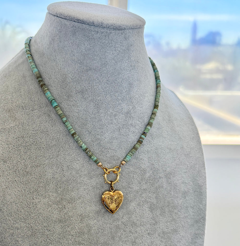A beaded necklace with turquoise and brown beads, gold-toned metal links, and a vintage Carl Art gold heart-shaped pendant, displayed on a gray fabric jewelry bust.