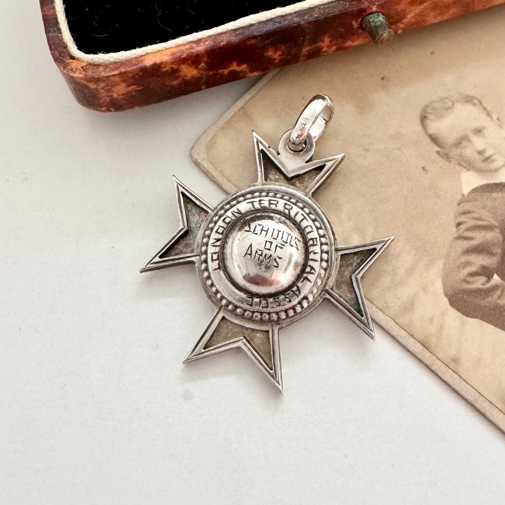 An antique sterling silver star-shaped watch fob with a circular center medallion engraved with 'SCHOOL OF ARMS' displayed on a vintage photograph next to an old wooden box.