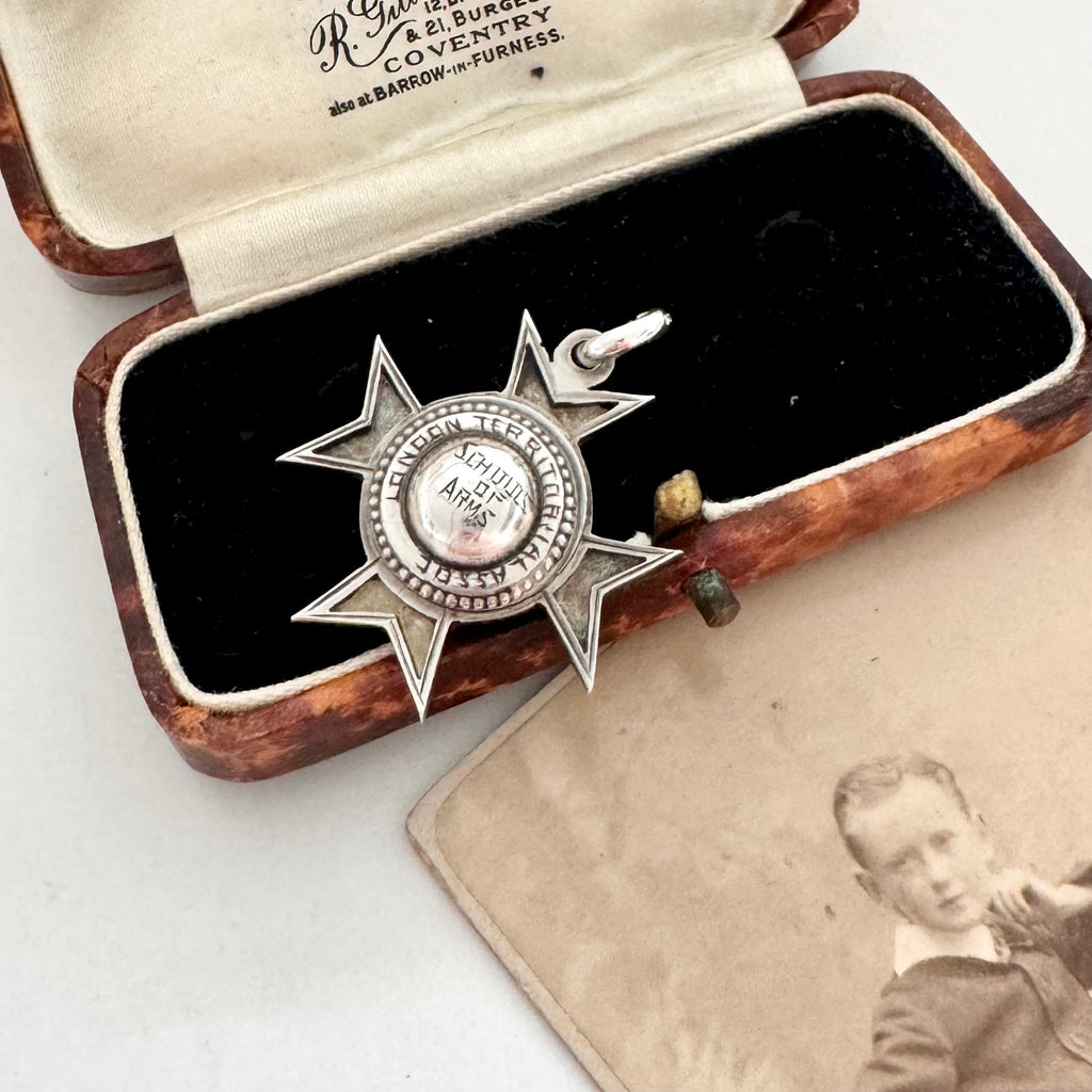 An antique sterling silver star-shaped watch fob with a circular center medallion engraved with 'SCHOOL OF ARMS' displayed on a vintage photograph next to an old wooden box.