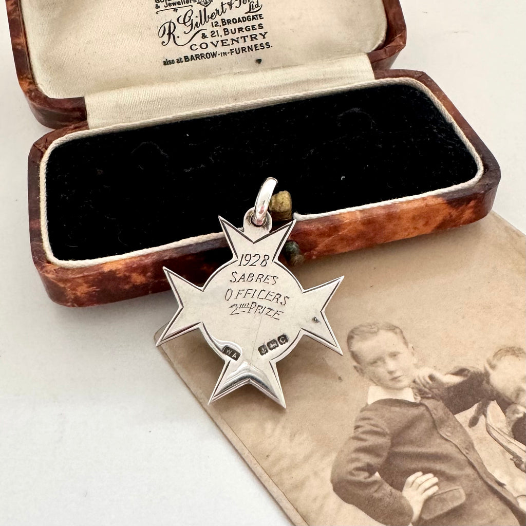 An antique sterling silver star-shaped watch fob with a circular center medallion engraved with 'SCHOOL OF ARMS' displayed on a vintage photograph next to an old wooden box.