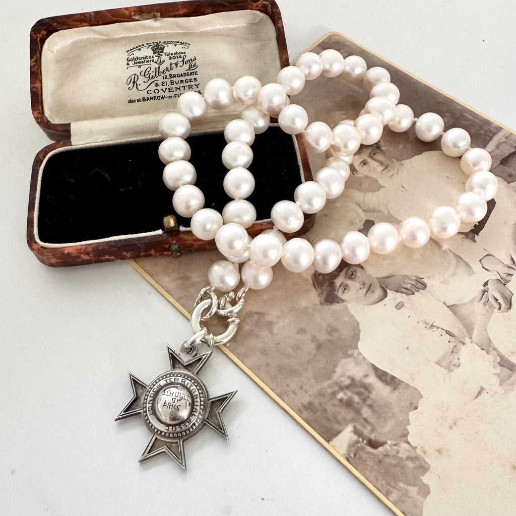 An antique sterling silver star-shaped watch fob with a circular center medallion engraved with 'SCHOOL OF ARMS' on a pearl necklace and displayed on a vintage photograph next to an old wooden box.