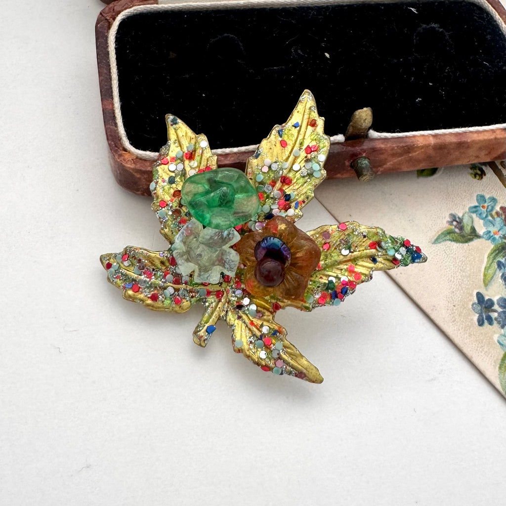 A vintage European hand wired gold-toned brooch shaped like a lotus flower with green, amber, and multicolor rhinestone accents, displayed near an old jewellery box and vintage postcard