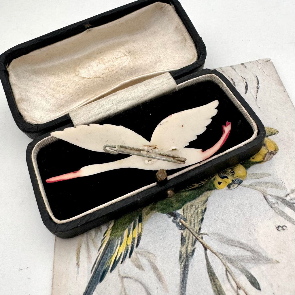 A vintage 1930's flying stork brooch or pin with cream-colored body and wings, black feather accents, and a red beak, displayed on a printed card with an open dark case in the background.