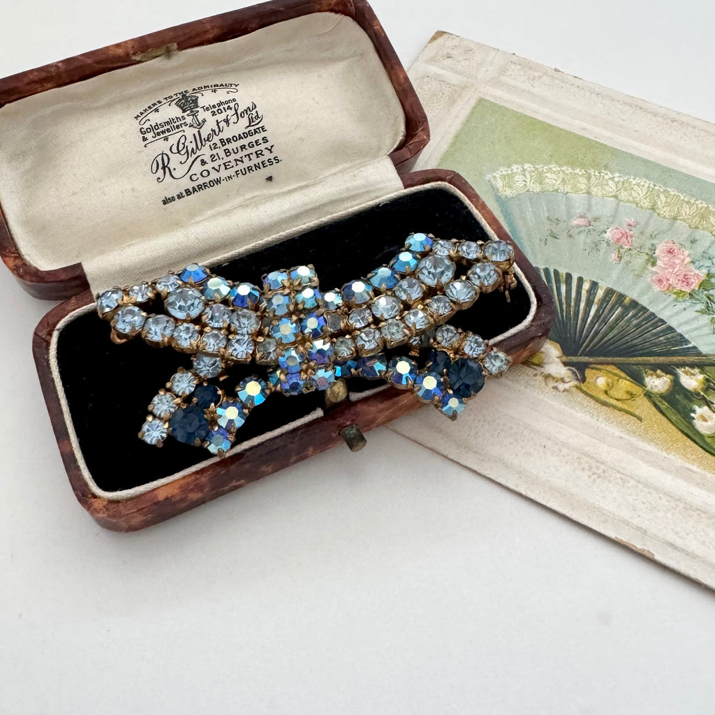 A vintage rhinestone brooch with blue and clear stones, displayed in an open wooden jewelry box next to a decorative fan illustration on a white background.