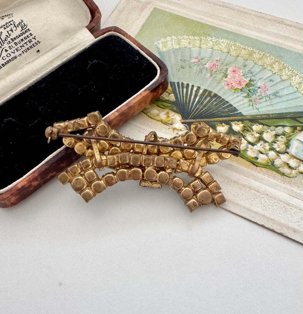 Back of a vintage rhinestone brooch, displayed next to an open antique jewelry box and a decorative fan illustration on a light surface.