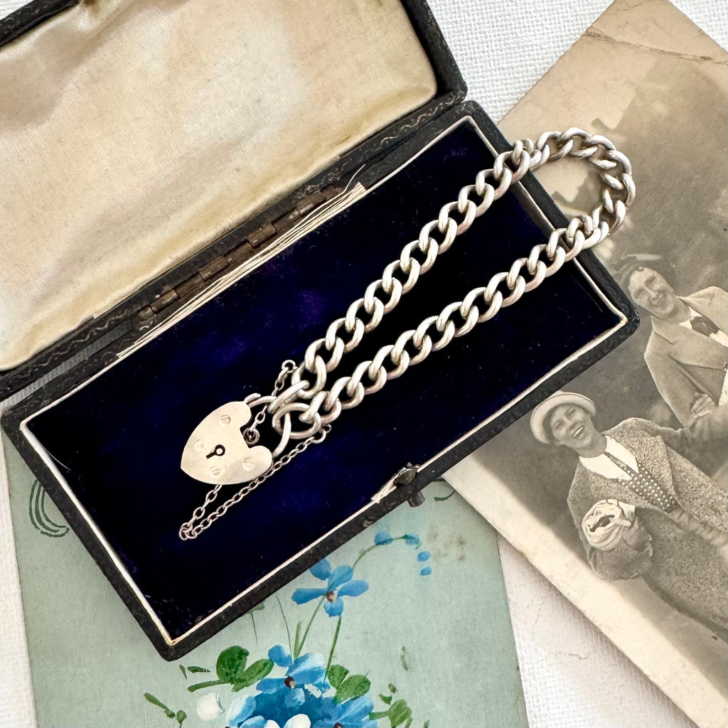 Silver chain bracelet with a heart padlock in an open velvet box on an old photograph background.