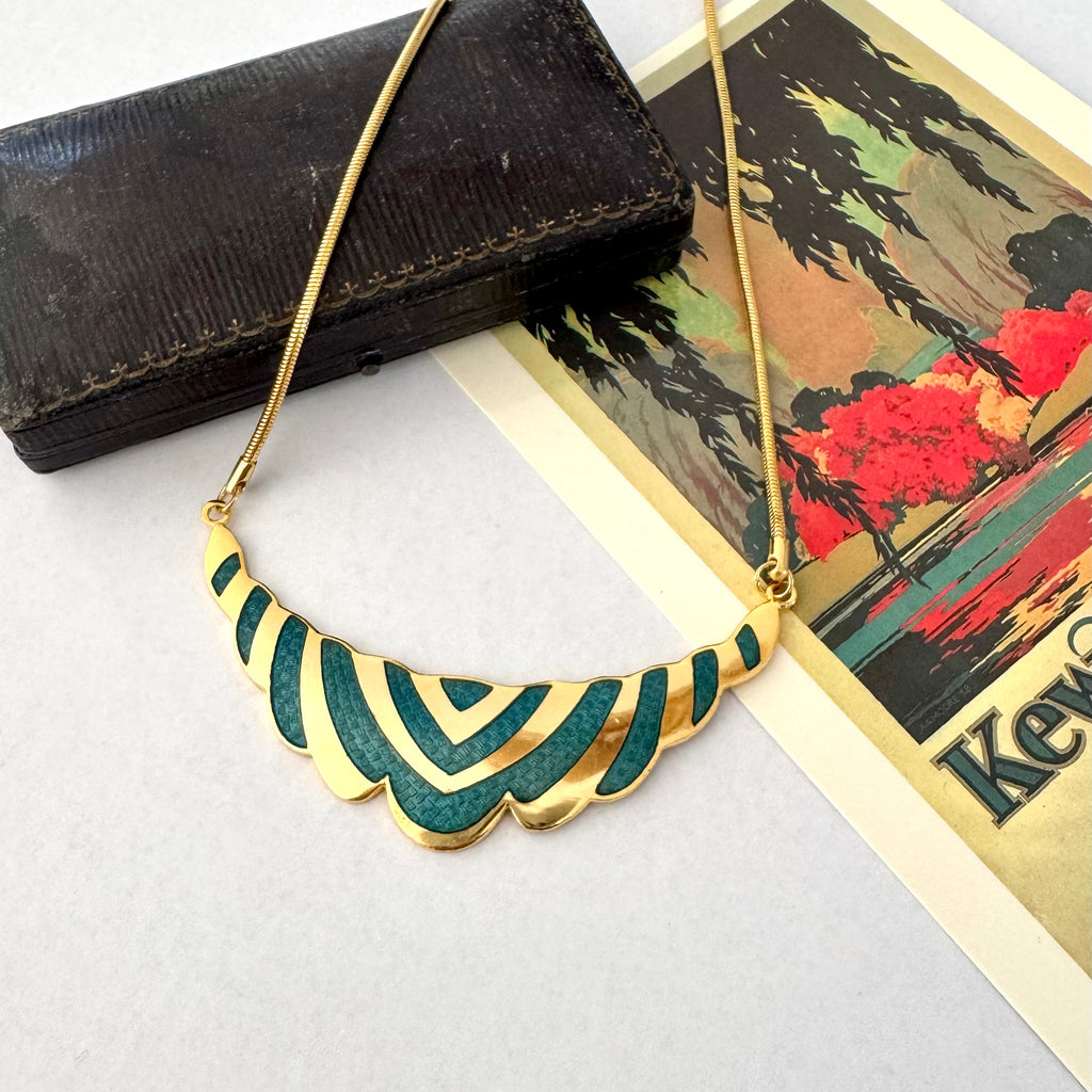 Gold necklace with a scalloped Art Deco style pendant featuring green inlay, displayed on a light gray surface next to a vintage landscape card and a dark textured box.