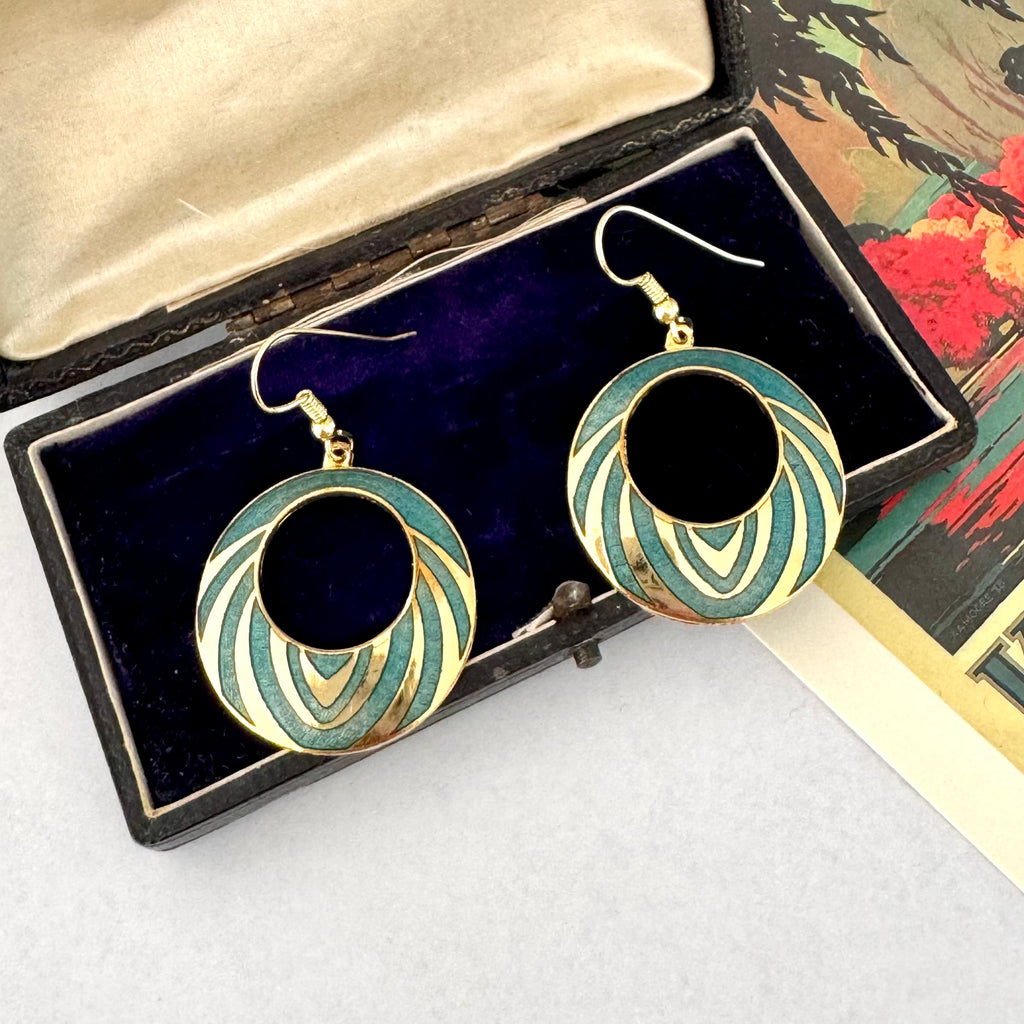 A pair of gold-tone hoop earrings with concentric circular enamel detailing in teal and cream, displayed inside an open black jewelry box with a dark purple velvet lining.