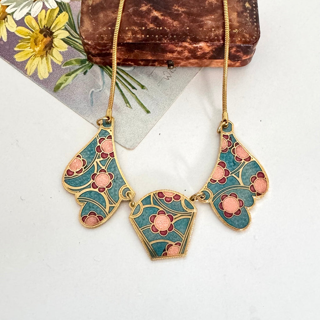A vintage 1980's gold-tone statement necklace with three teardrop-shaped enamel pendants featuring a teal, pink, and gold floral pattern, displayed on a white surface with a vintage card and brown box as props.