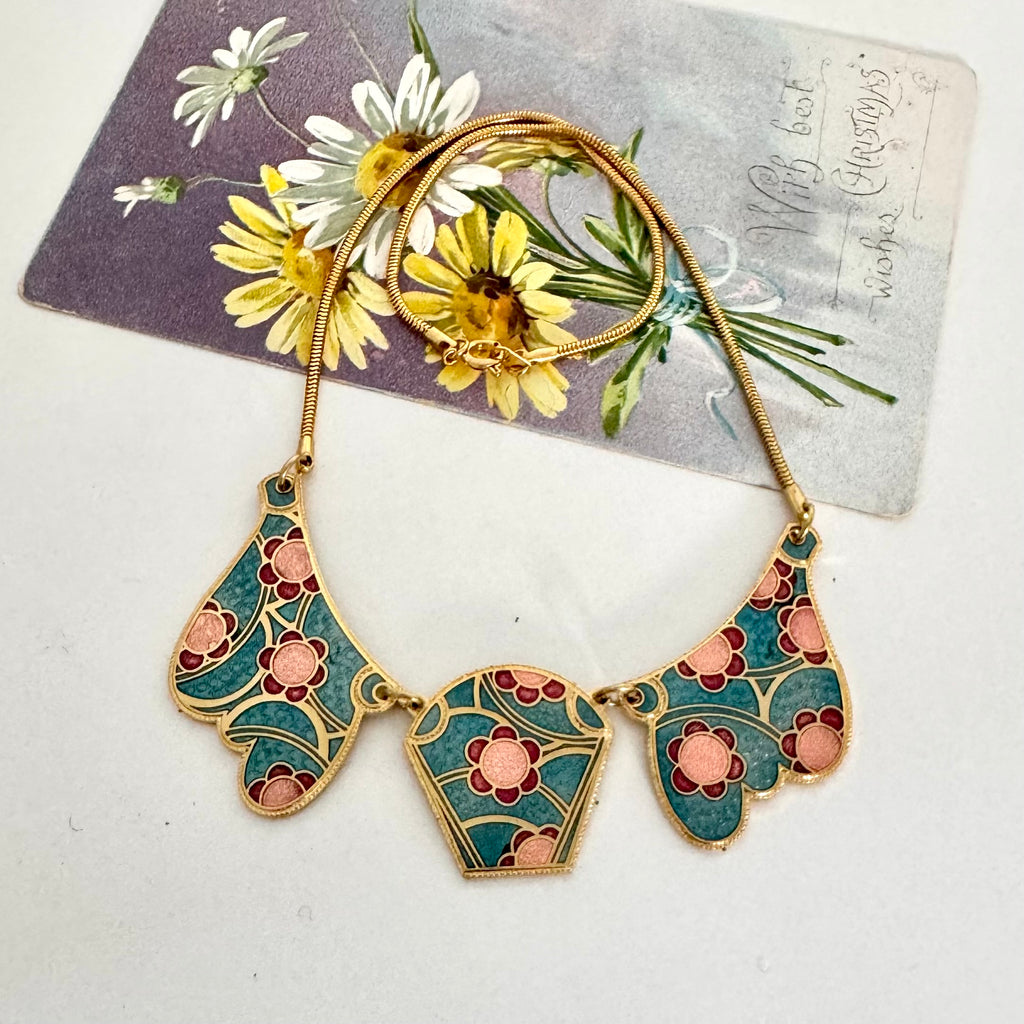 An Art Deco style gold-plated necklace with three teardrop-shaped enamel pendants featuring teal, pink, and gold floral patterns, displayed on a vintage floral postcard.