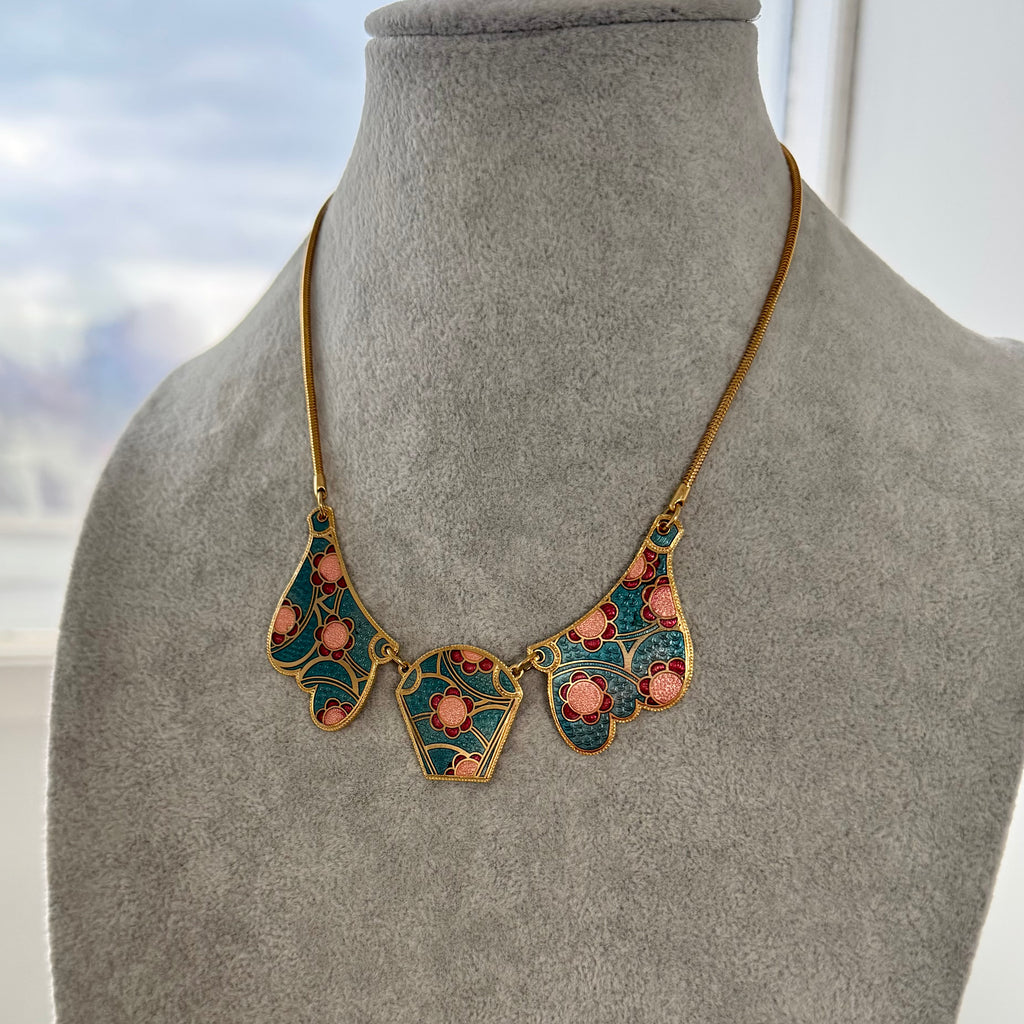 A vintage 1980's ornate Art Deco style necklace displayed on a gray velvet jewelry bust. The necklace features a gold-tone chain and a central multi-piece bib pendant with teal enamel, gold filigree, and pink floral accents.