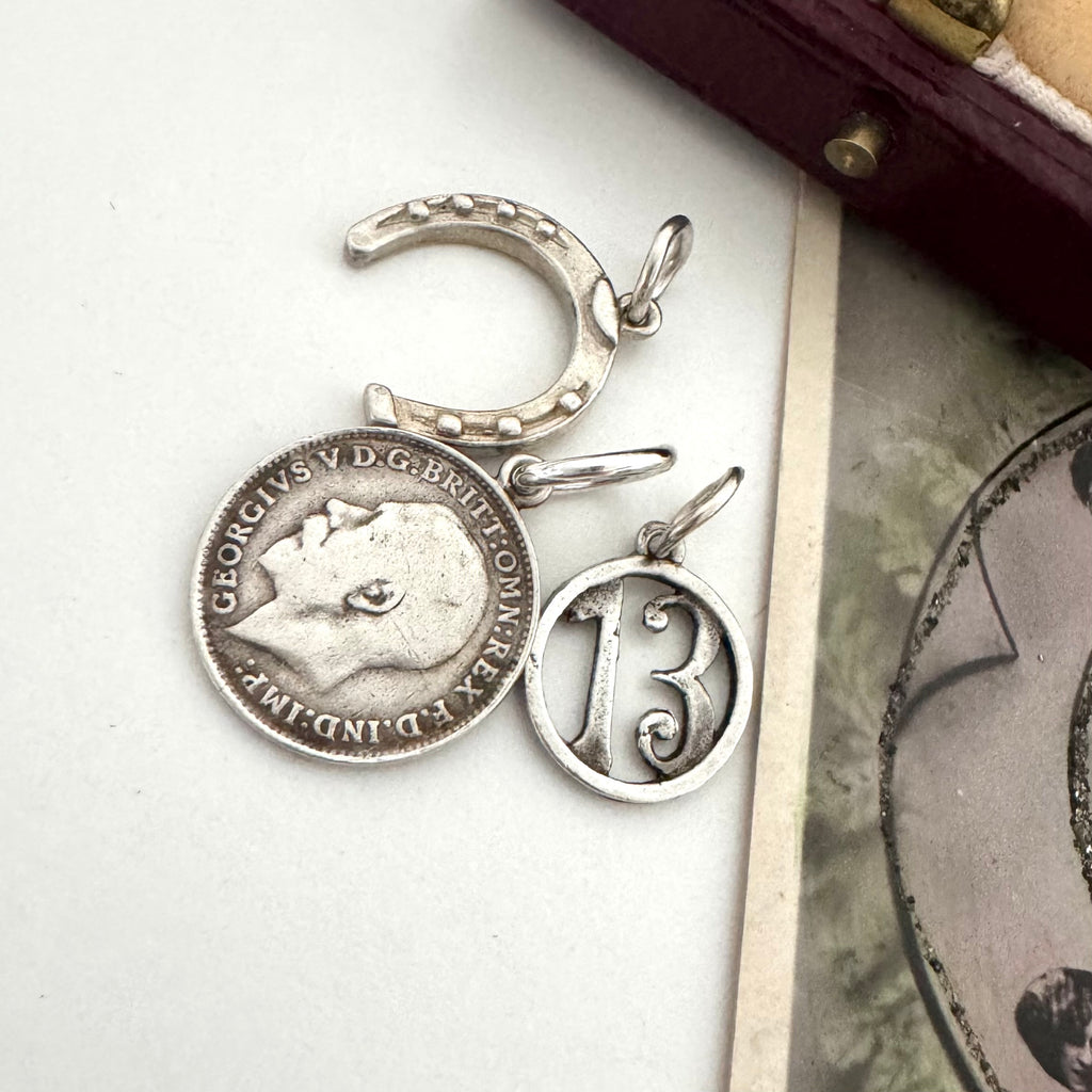 A vintage silver horseshoe charm, no 13 charm and a 1918 British threepence on a white background with an old postcard in the corner.