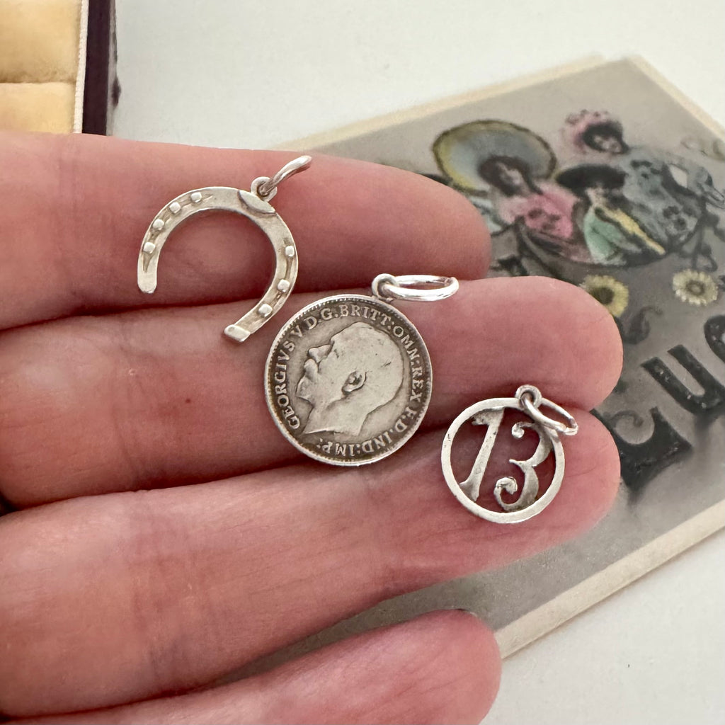 A vintage silver horseshoe charm, no 13 charm and a 1918 British threepence held in a hand with an old postcard in the corner.