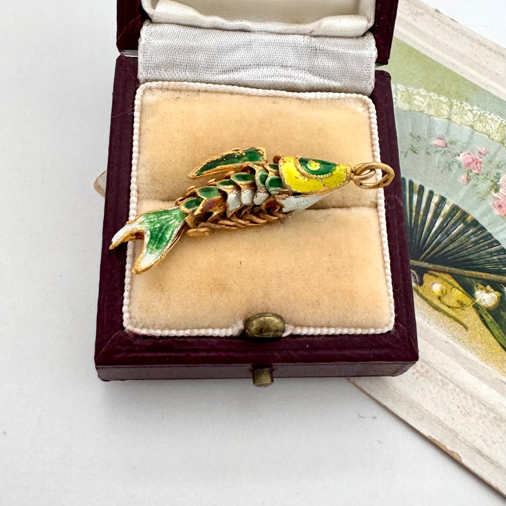 Vintage articulated green and yellow fish charm in a box with a vintage postcard in the background