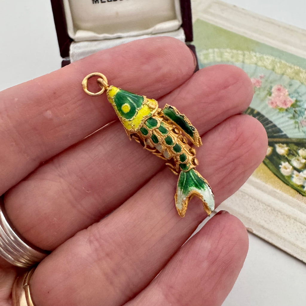 Vintage articulated fish charm in someones hand and with a box and a vintage postcard in the background