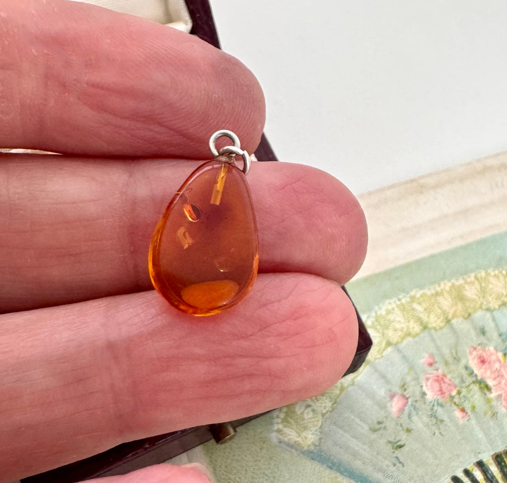 Teardrop-shaped amber pendant held between fingers with a floral-patterned fabric background