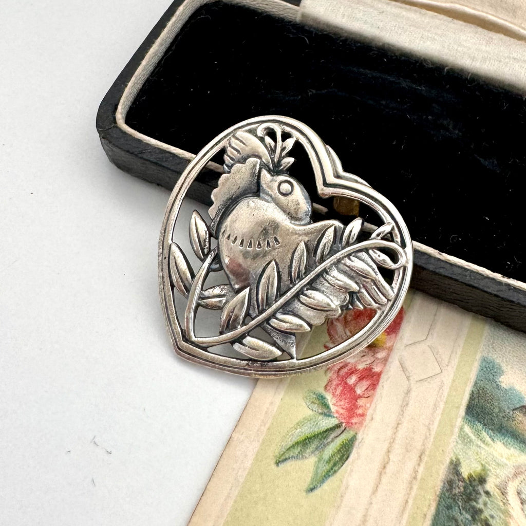 Silver bird and leaf design on a heart-shaped pendant with a black box in the background.