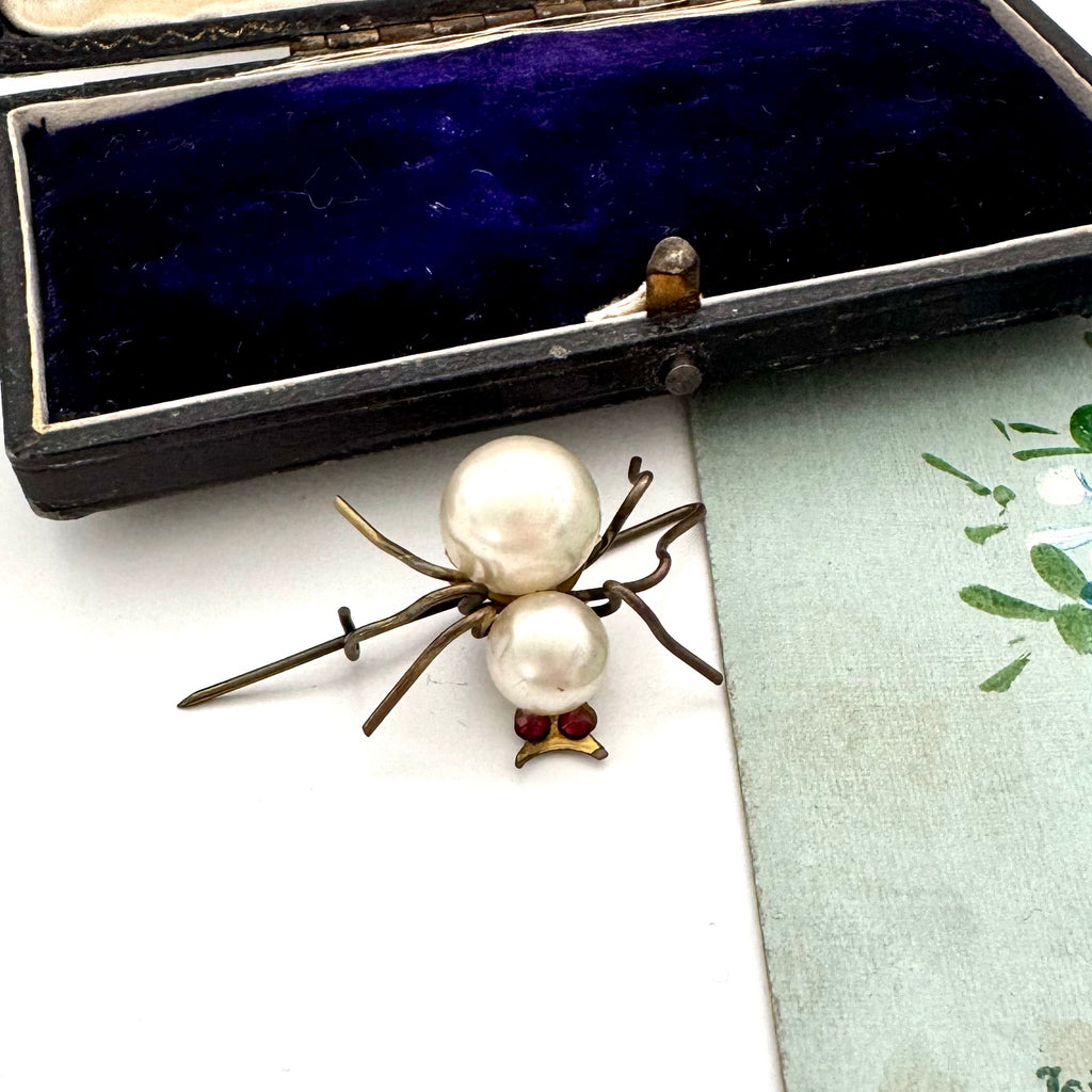Spider-shaped brooch with pearls on a white surface next to an open jewelry box.