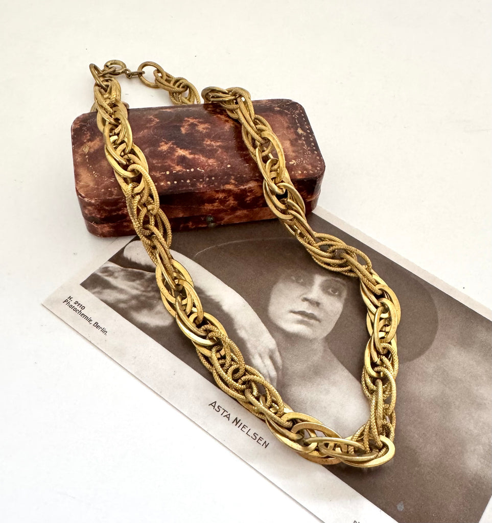 A chunky French vintage rolled gold chain necklace draped over a vintage brown textured box and a black-and-white print featuring the name 'ASTA NIELSEN'.