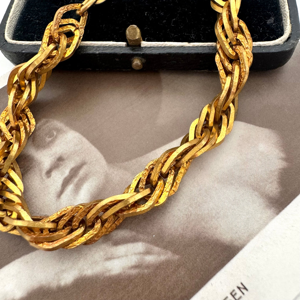 A chunky textured gold chain bracelet draped over a black jewelry box, with a vintage black-and-white portrait print underneath.