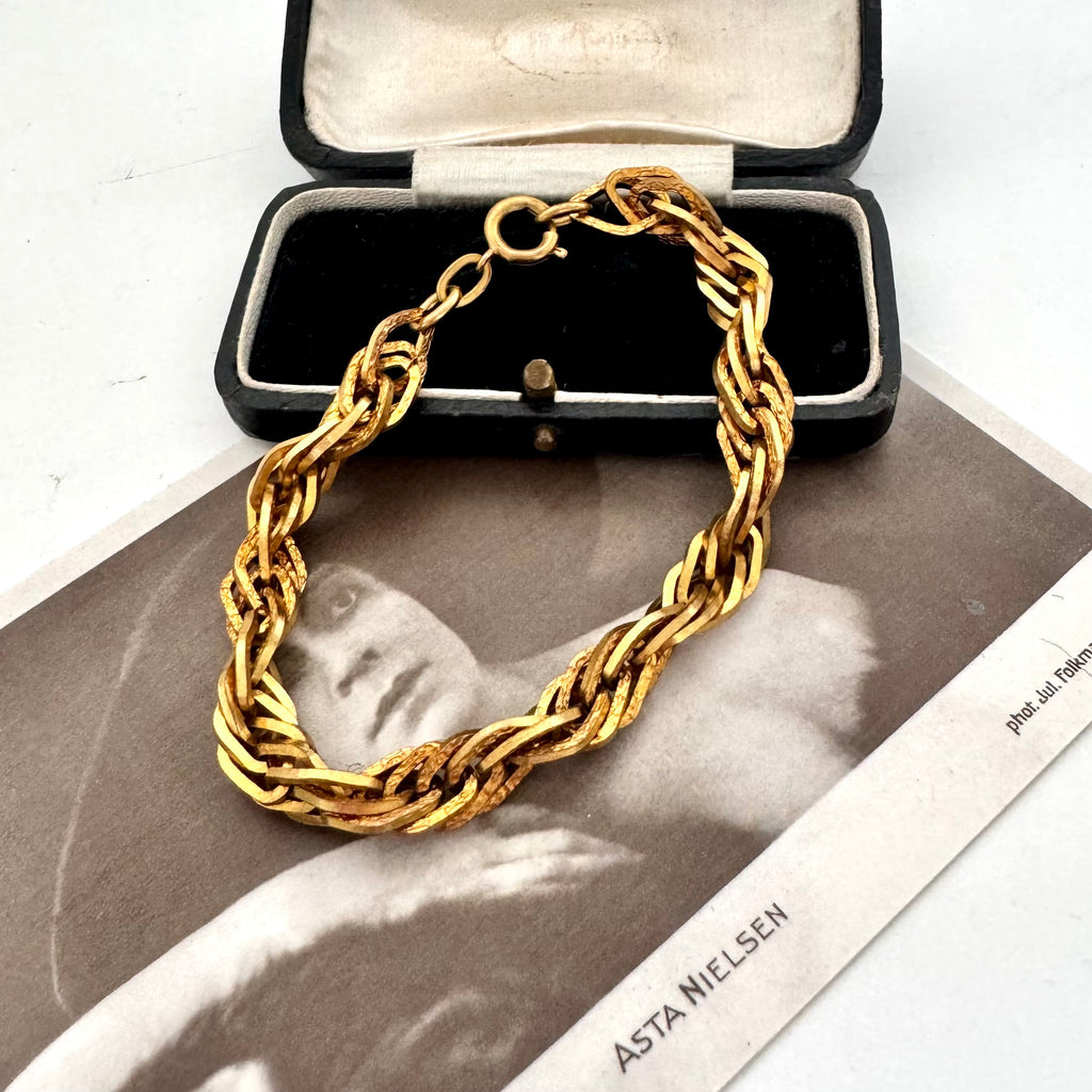A chunky gold chain link bracelet displayed on top of an open black jewelry box, with a vintage black-and-white photograph underneath.