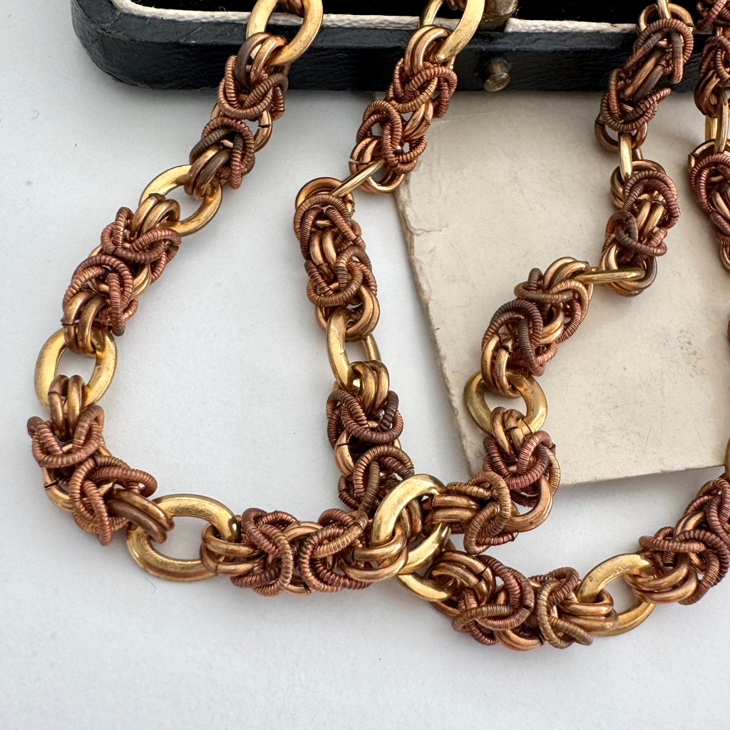 A close up of a vintage-Jakob Bengel Byzantine chain necklace with interwoven copper and gold links, draped over a black leather case and an old sepia-toned family photograph on a white surface.