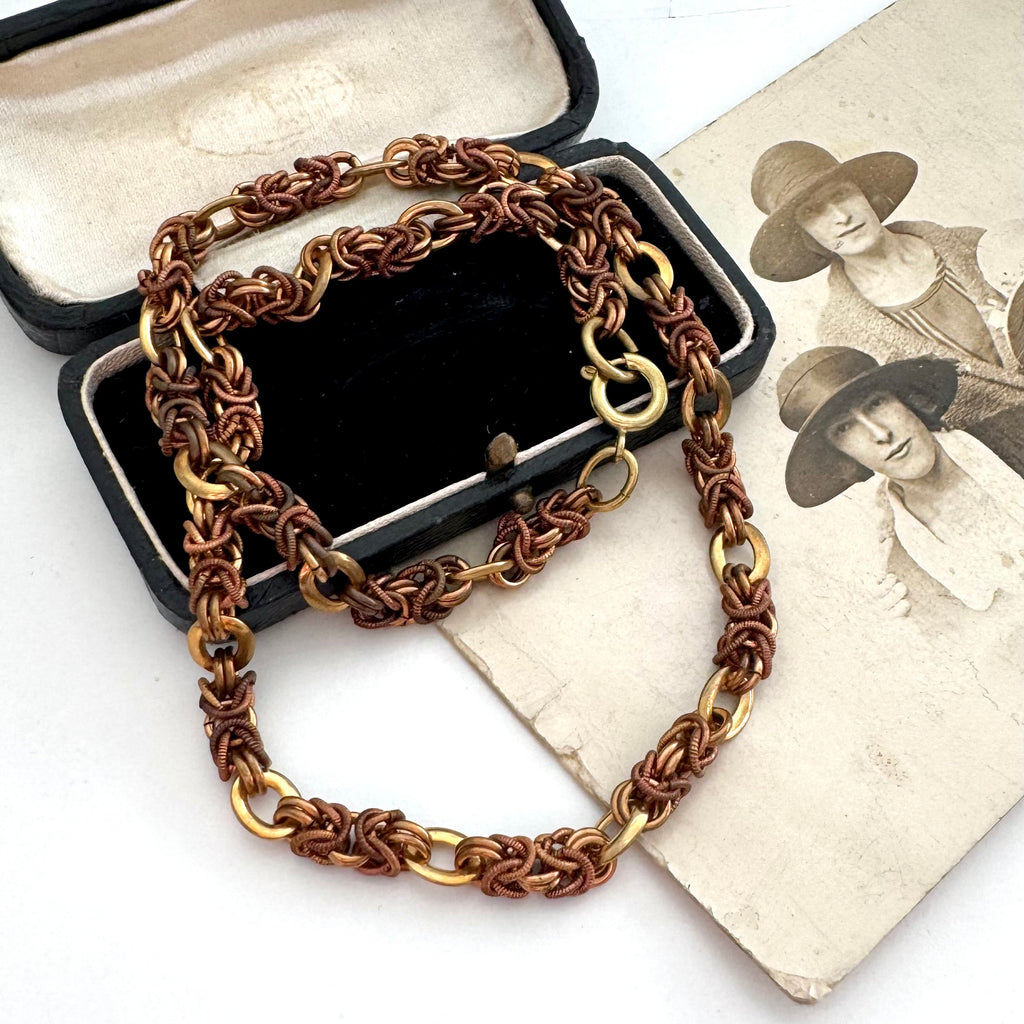 A vintage-Jakob Bengel Byzantine chain necklace with interwoven copper and gold links, draped over a black leather case and an old sepia-toned family photograph on a white surface.