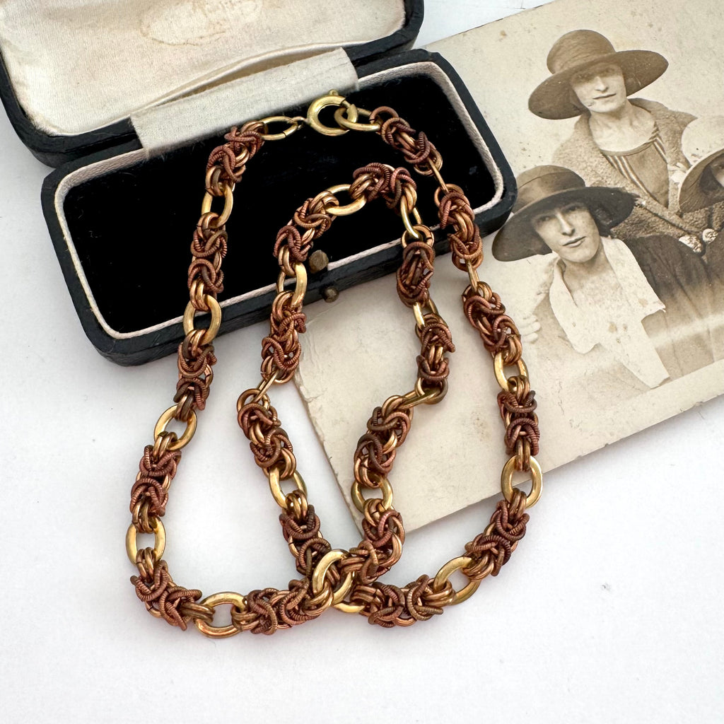A vintage-Jakob Bengel Byzantine chain necklace with interwoven copper and gold links, draped over a black leather case and an old sepia-toned family photograph on a white surface.
