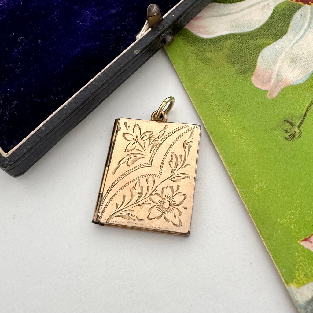 Gold engraved locket on a white surface with a floral card and black box in the background