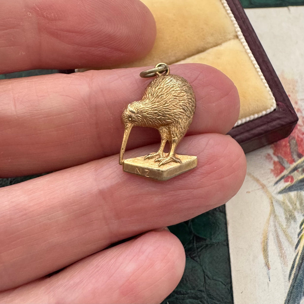 Gold kiwi bird pendant held between fingers with a textured background