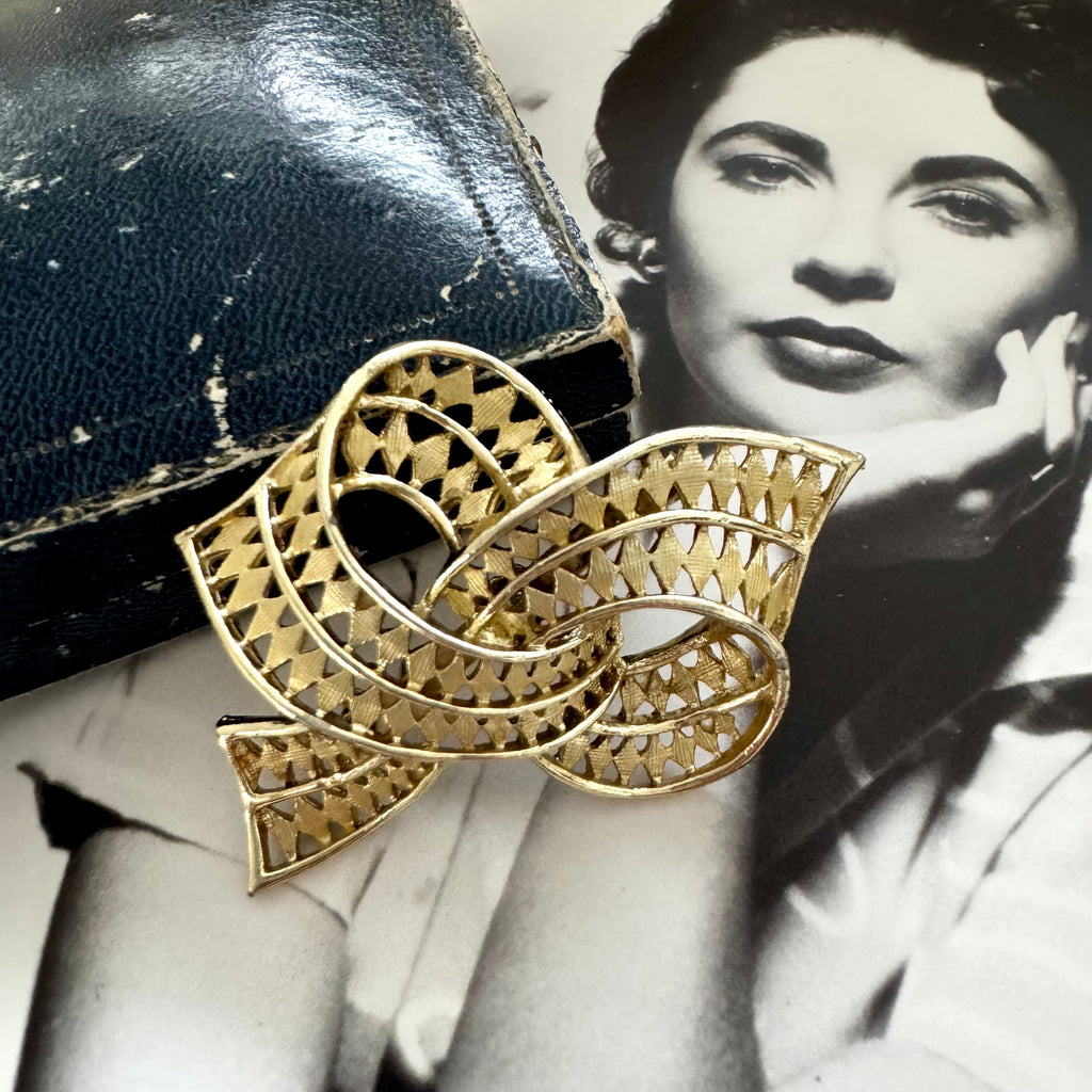 Vintage gold tone bow brooch with intricate design on a black and white photo of Elizabeth Taylor in the background