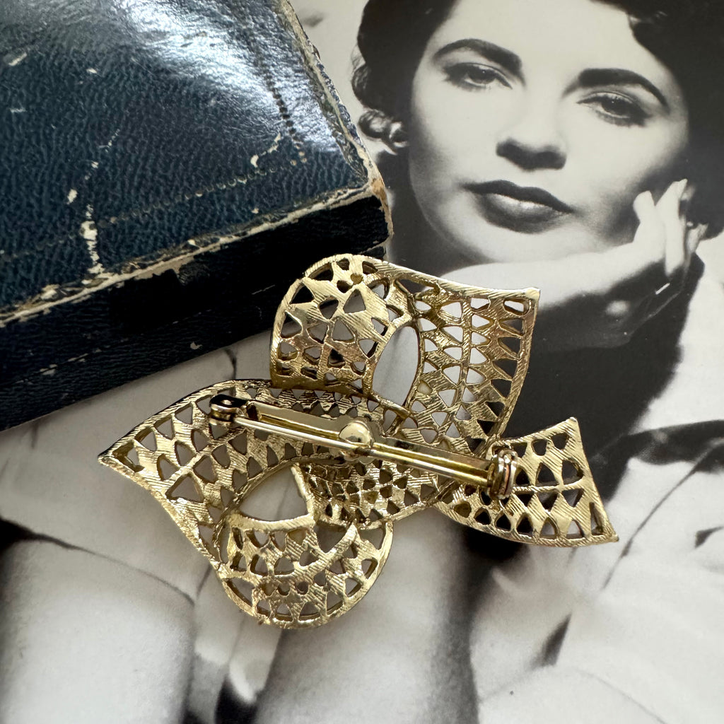 Gold bow-shaped brooch on a black leather surface with a vintage-style portrait in the background.