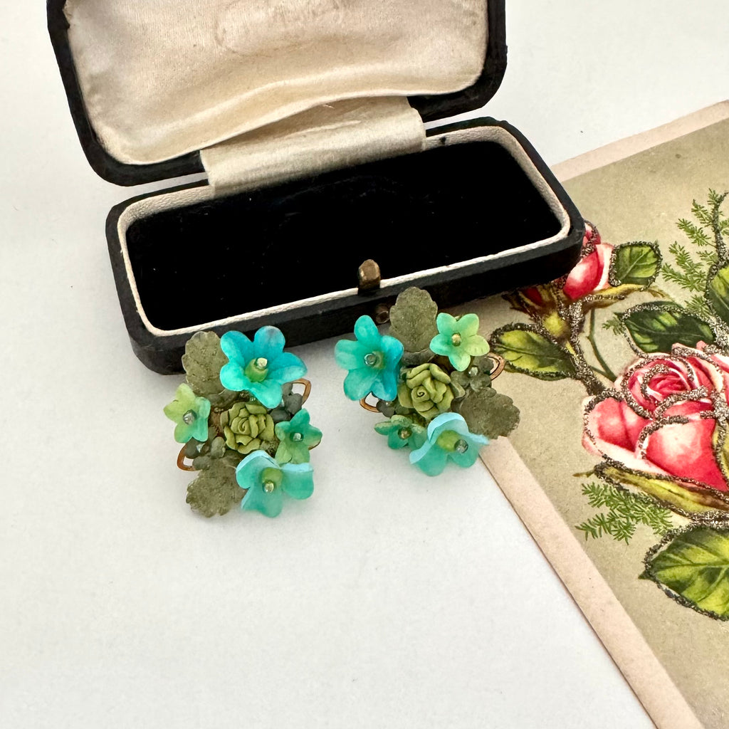 Floral vintage earrings with green and blue flowers in an open jewelry box on a white surface.
