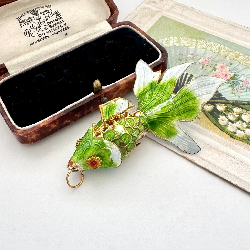 A green and white enamel koi fish pendant with gold-tone detailing, displayed next to a vintage presentation box on a white surface.