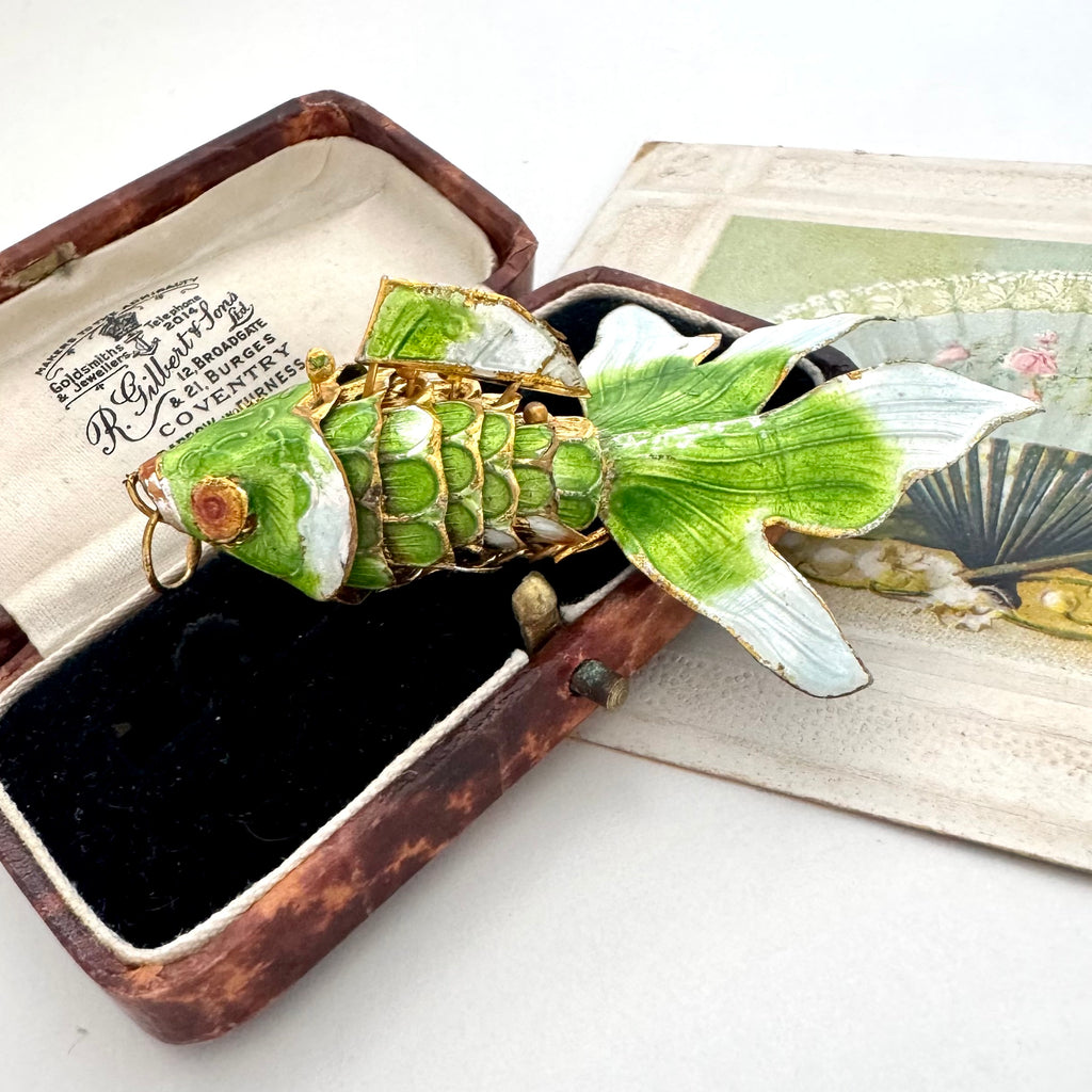 An vintage koi fish-shaped pendant with green and white enamel scales and gold wire detailing, displayed inside an open vintage brown presentation box with a black velvet lining.