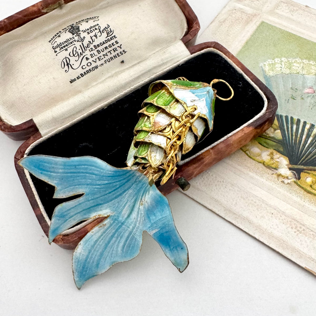 A decorative fish-shaped pendant with a blue tail and green-and-white scaled body, displayed in an open vintage jewelry box. The box lid shows printed branding text, and part of a fan illustration is visible on the right.