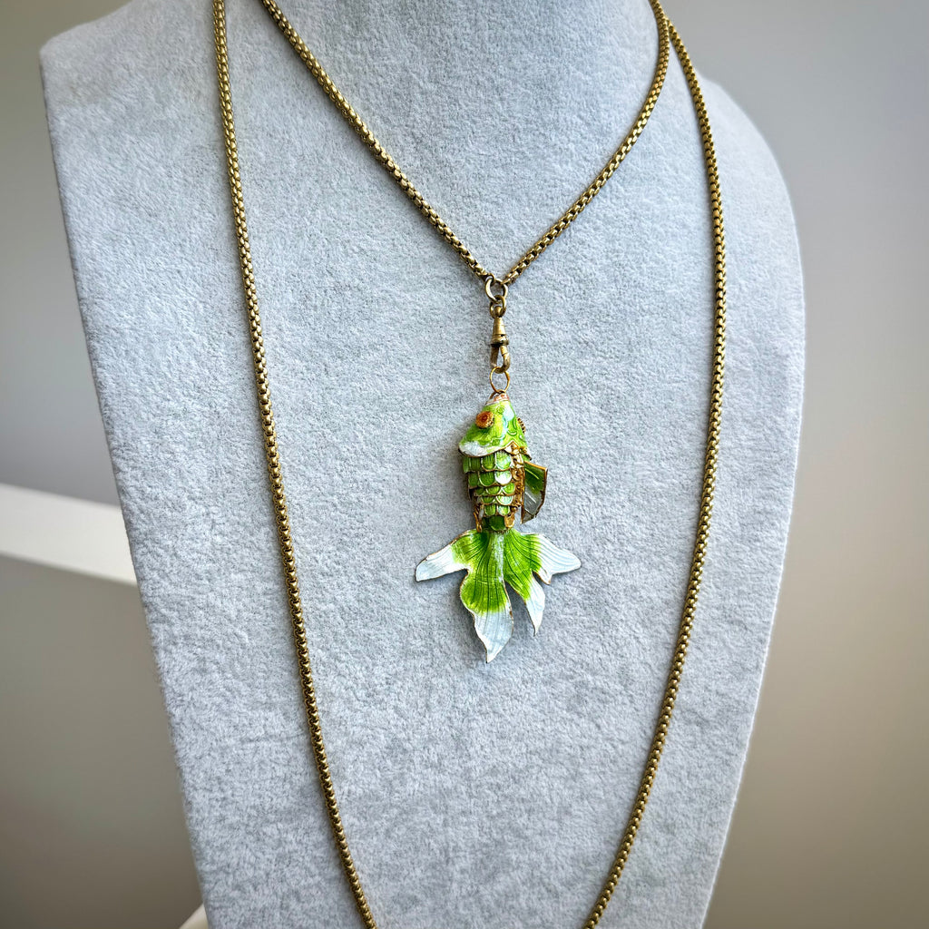 A gold chain necklace with a fish-shaped pendant featuring green, white, and gold enamel details, displayed on a gray fabric jewelry bust.